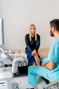 Ameliyattan sonra bir kız hasta ultrason teşhis odasında sedyede oturur ve bir doktoru dikkatle dinler.