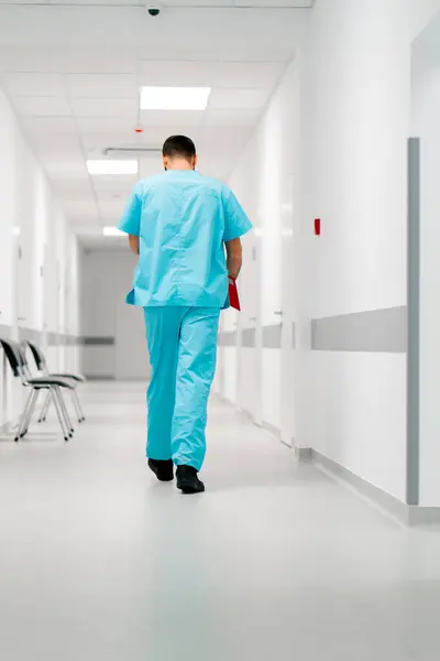 A tall male doctor with a beard walks along the hospital corridor with a folder of documents