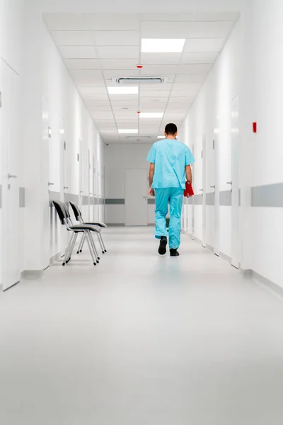 A tall male doctor with a beard walks along the hospital corridor with a folder of documents