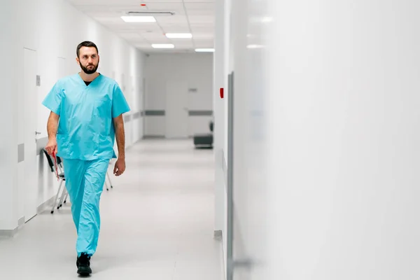 A tall male doctor with a beard walks along the hospital corridor with a folder of documents and enters his office