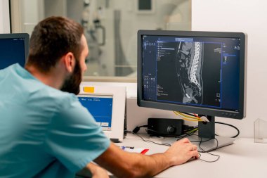 Bir radyolog bilgisayar monitörünün arkasındaki masada oturur ve bir hastanın muayenesi sırasında manyetik rezonans görüntüleme görüntüsünü inceler.