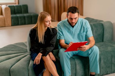 Doktor koridorda hastasıyla birlikte kanepede oturur ve test sonuçlarına göre ona tedavisini anlatır.