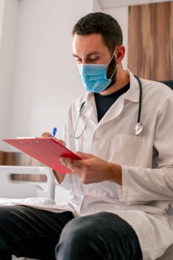 Tıbbi maske ve hastane önlüğü takmış bir doktorun tıbbi geçmişine hastanın şikayetlerini yazdığı yakın plan bir fotoğraf.