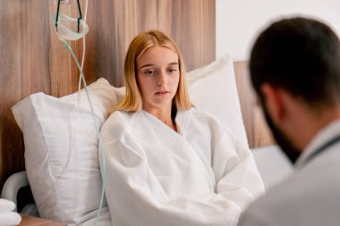 Hasta bir kız hastane odasında uzanıyor ve göğsünü eliyle tutuyor ve doktorun yanında öksürüyor.