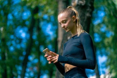 Spor kıyafetli bir kızın yeşil bir parkta antrenman yaparken telefonuna dikkatlice bakarken çekilmiş bir fotoğrafı.