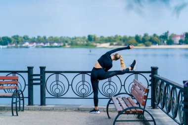 Genç bir sporcu kız sabah nehir kıyısındaki sette bacak esnekliği için esneme hareketleri yapıyor.