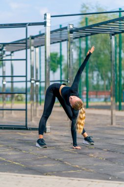 Genç bir bayan sporcu açık hava spor sahasında esneklik ve sırt sağlığı için ısınıyor.