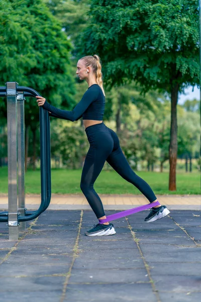 Focused girl athlete performs exercises with a sports elastic band on street exercise machines