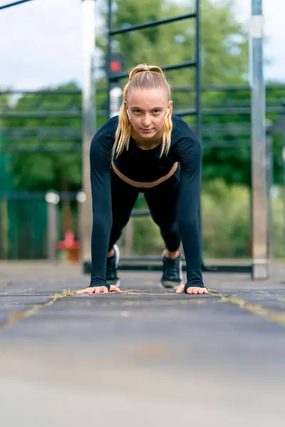 On a sports street playground a pumped-up girl stands in a plank and listens to motivating music on headphones