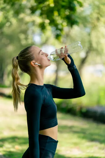 Athletic young woman after an active morning jog and workout drinks water from a bottle on the alley in the park