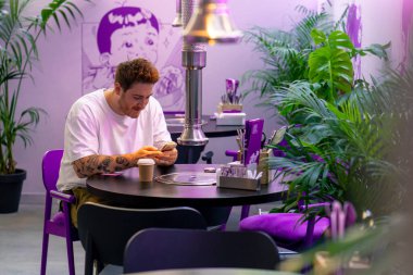 Genç bir adam kafeteryada bir masada oturuyor. Güzel bir iç mekanda. Elinde akıllı telefonuyla siparişini bekliyor.