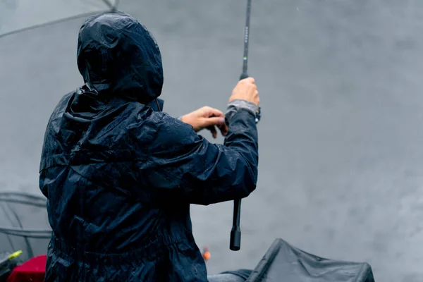Fisherman in raincoat with fishing rod or spinning professional tools sitting on river bank rear view Pull the fish out of the lake sport fishing