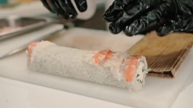 Siyah eldivenli bir suşi makinesinin yakın çekimi. Mutfakta California usulü rulo hazırlarken bıçakla karides filetosu kesiyor.