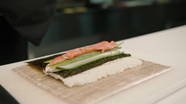一名戴着黑色手套的寿司制造商在厨房桌上用米片鲑鱼片凝乳芝士黄瓜鳄梨制作寿司的特写镜头 — 图库视频影像