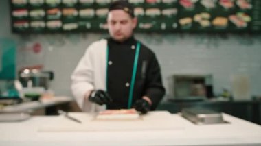Suşi şefi, beyaz mutfak tahtasında somon ve salatalıklı krem peynir hazırlıyor.