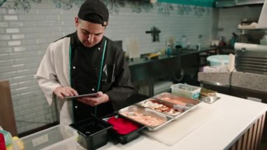 Elinde tabletle dalgın bir suşi şefi lezzetli böreklerin tariflerine bakar ve malzemelerini seçer.