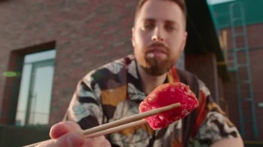 Elinde çubuklarla terasta oturmuş suşi yiyen genç ve çekici bir adamın yakın çekimi.