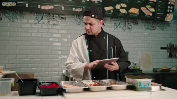 Egy Töprengő Sushi Séf Tablettával Kezében Megnézi Ízletes Tekercsek Receptjeit — Stock videók