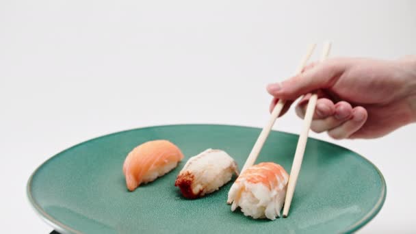 箸を使って男性の手を閉じ 白い背景にサーモンとウナギの寿司のプレートからエビ寿司を1つ取る — ストック動画