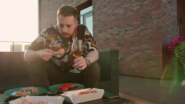 テラスデッキに座っている寿司ロールを食べる手のワインのグラスを持つ若い魅力的な男 — ストック動画