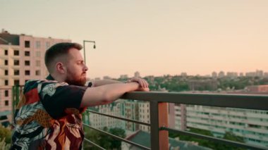 Genç sakallı bir adam terasta dikiliyor. Geleceği düşünerek şehre bakıyor.