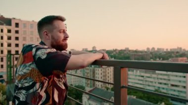 Genç sakallı bir adam terasta dikiliyor. Geleceği düşünerek şehre bakıyor.