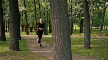 Spor giyim ve kulaklık giyen genç bir kız sabahları parkta koşuyor. Fiziksel olarak aktif yaşam tarzını sürdürüyor.