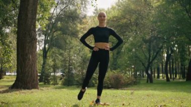 Sabah sporu yapan bir kız temiz havada egzersiz yaparken bacağı kalçasını esnetirken dairesel hareketler yapıyor.