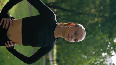Spor giyim ve kulaklık giyen bir kızın dikey video portresi sabah egzersizi ve parkta antrenman yaptıktan sonra yoruldu.