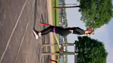 Spor üniformalı güzel kız spor caddesindeki oyun parkında bacaklarını lastik bantla şişirmek için egzersiz yapıyor.