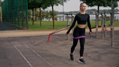 Spor üniformalı güzel bir kız spor sahasında bacaklarını lastik bantla şişirmek için egzersiz yapıyor.