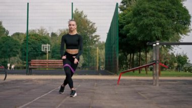 Spor üniformalı güzel bir kız spor sahasında bacaklarını lastik bantla şişirmek için egzersiz yapıyor.