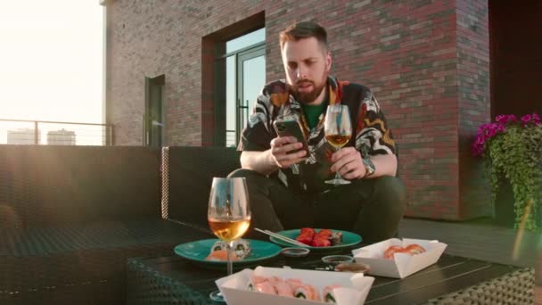 年轻迷人的男人 手里拿着杯酒 坐在平台上吃寿司卷 — 图库视频影像