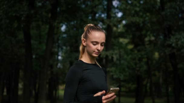 一个穿着运动服的女孩在公园的大街上一边认真地看着一边打电话 选择在线训练 — 图库视频影像