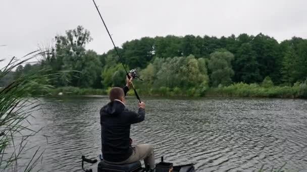 釣りロッドまたはスピニングと川岸の背面ビューに座っているプロのツールを持つ漁師 湖のスポーツ釣りから魚を引っ張る — ストック動画