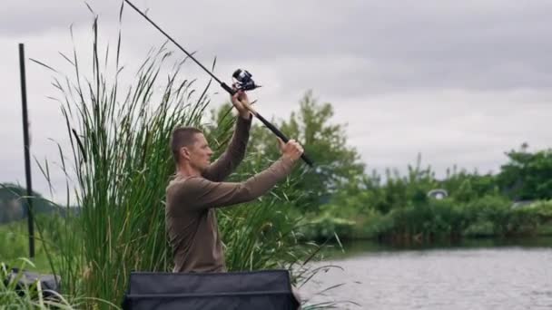 釣り棒または回転および湖のスポーツの釣りから川岸に座っている専門用具が付いている漁師 — ストック動画