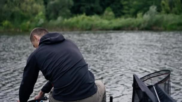 釣りロッドまたはスピニングと川岸の背面ビューに座っているプロのツールを持つ漁師 湖のスポーツ釣りから魚を引っ張る — ストック動画