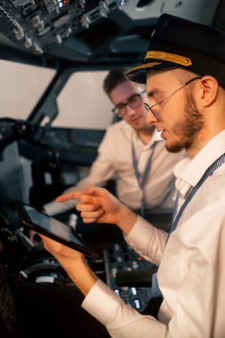 Pilotlar kontrol panelinin yanındaki kokpitte ellerinde tabletle uçuş simülatörü başlamadan önce rotayı tartışıyorlar.