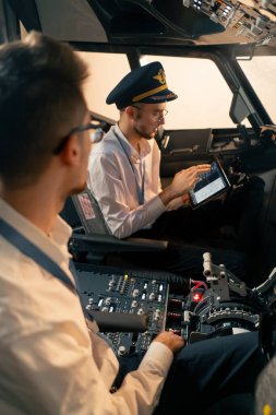 Pilotlar kontrol panelinin yanındaki kokpitte ellerinde tabletle uçuş simülatörü başlamadan önce rotayı tartışıyorlar.