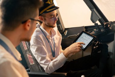 Pilotlar kontrol panelinin yanındaki kokpitte ellerinde tabletle uçuş simülatörü başlamadan önce rotayı tartışıyorlar.