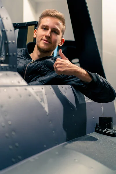 satisfied young guy sitting in flight simulator of military plane after virtual flight shows hand sign super fun