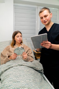 Estetik ameliyattan sonra hastanın odasındaki doktor elinde bir tablet tutuyor. Kız aynaya bakıyor ve sonucu değerlendiriyor.