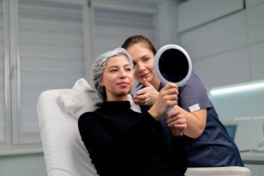 Güzellik salonundaki güzel bayan müşteri aynaya bakıp doktorla birlikte estetik ameliyatın sonucunu kontrol ediyor.