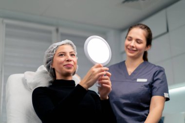 Güzellik salonundaki güzel bayan müşteri aynaya bakıp doktorla birlikte estetik ameliyatın sonucunu kontrol ediyor.
