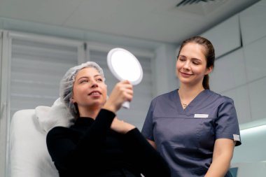 Güzellik salonundaki güzel bayan müşteri aynaya bakıp doktorla birlikte estetik ameliyatın sonucunu kontrol ediyor.