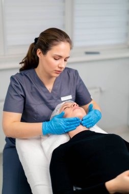 Eldivenli kozmetik uzmanı, gençleştirme işlemi sırasında müşterinin yüzüne ve boynuna elleriyle masaj yapıyor.