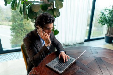 Ciddi bir adamın portresi. Ofiste dizüstü bilgisayarda çalışan bir iş adamı. İş arkadaşlarıyla akıllı telefondan iletişim kuruyor.