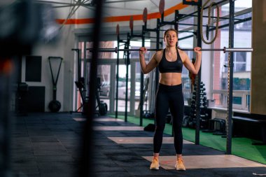 Spor salonunda, siyah bluzlu bir kız antrenör halterle egzersiz yapıyor.