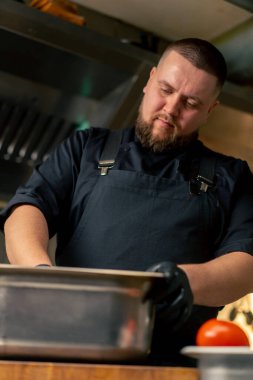 Profesyonel bir mutfakta, siyah ceketli bir aşçıda marinayla sebzeleri demir kasede karıştırıyor.