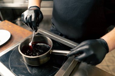 Siyah ceketli profesyonel bir aşçıya yakın sosu tencerede karıştırıyor.